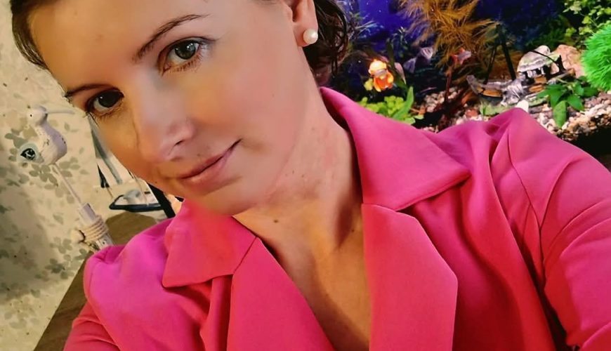 A woman with fair skin and light brown hair, wearing a bright pink blazer, takes a selfie in front of a fish tank filled with plants and decorations. The background features a patterned wall.