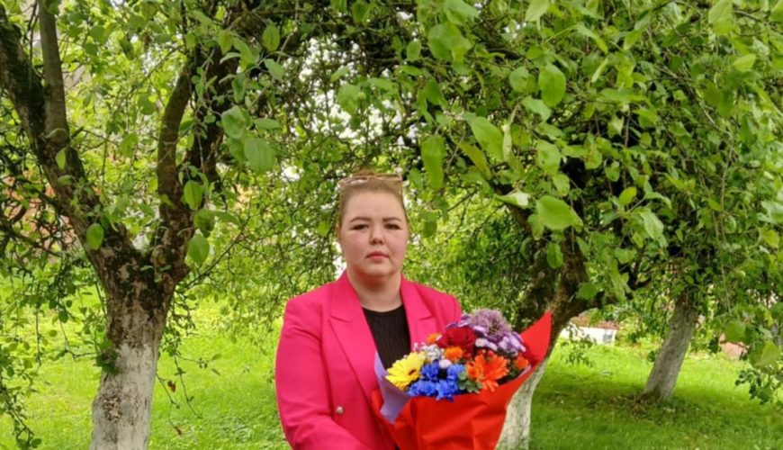 A person in a bright pink blazer and gray pants stands on green grass, holding a colorful bouquet of flowers, with leafy trees and lush greenery in the background.