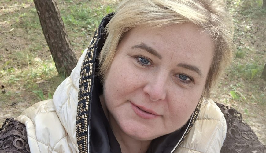A woman with blonde hair, wearing a light-colored puffer jacket with a hood, stands outdoors in a sunlit forest with trees and green grass visible in the background. She is looking at the camera and smiling slightly.