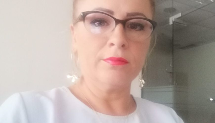 A woman with blonde hair in a bun, wearing glasses, red lipstick, a white top, and a necklace, sits indoors in an office setting with glass walls and a ceiling with lights.
