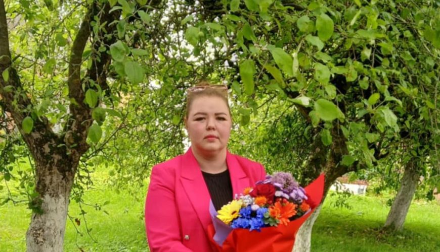 A person in a bright pink blazer stands on green grass under leafy trees, holding a colorful bouquet of flowers wrapped in red paper.