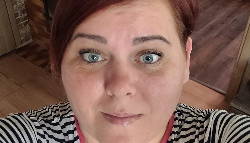 A person with short auburn hair and blue eyes takes a selfie indoors, wearing a black and white striped shirt with a pink collar. Wooden flooring and a doorway are visible in the background.