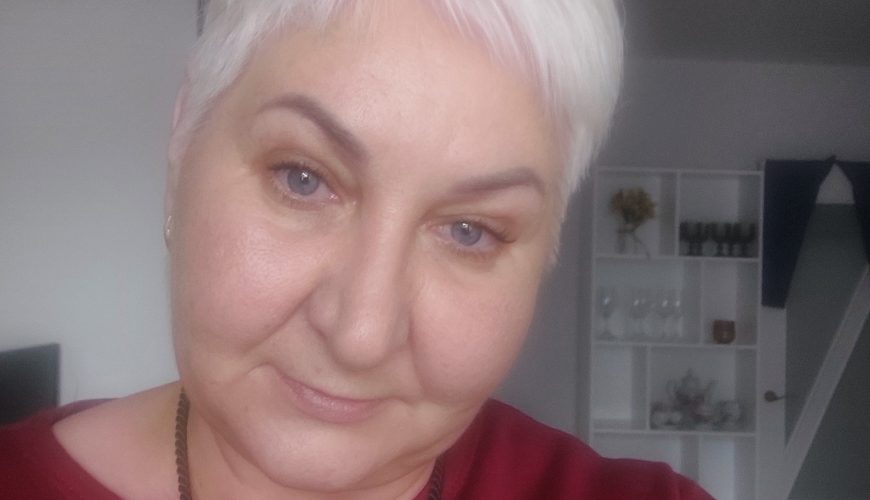 A woman with short white hair and a maroon top smiles softly at the camera. She is indoors, with shelves and a chandelier visible in the background.
