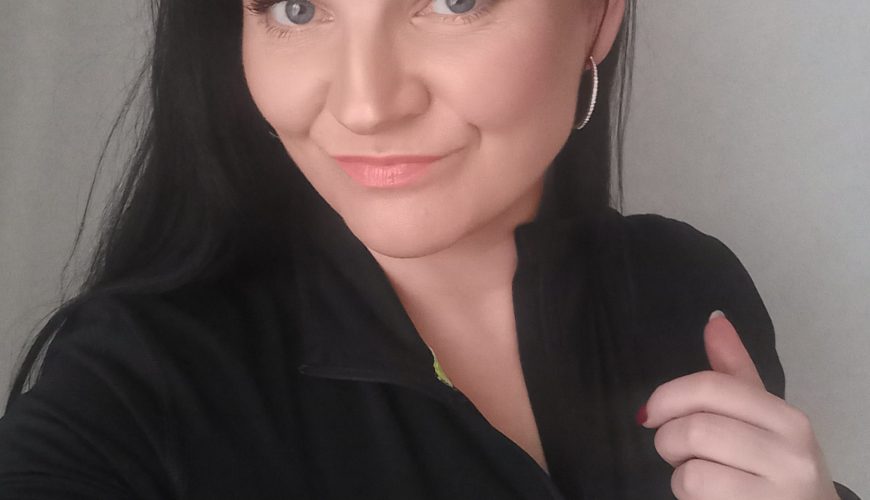 A woman with long dark hair and light eyes smiles at the camera, wearing a black zip-up top and hoop earrings, holding the collar of her top with one hand against a plain background.