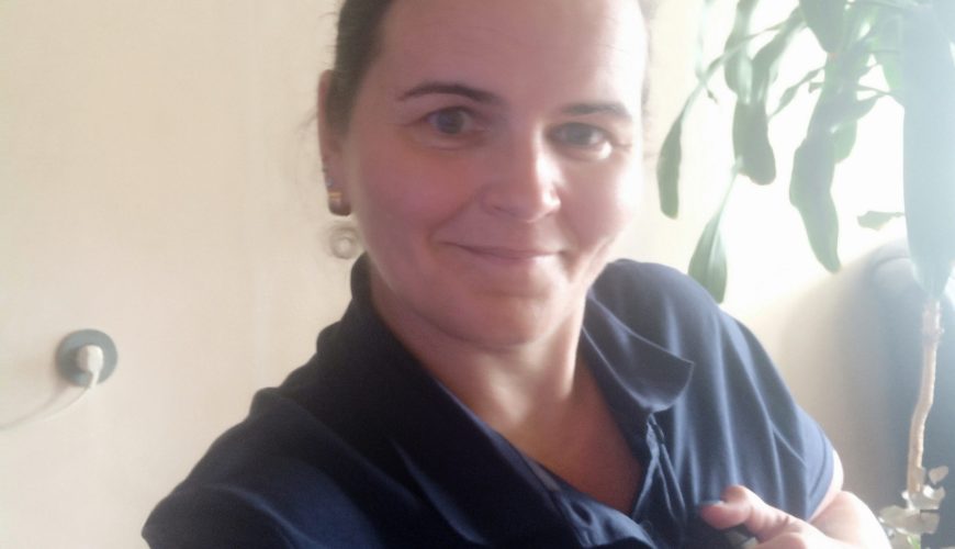 A woman smiles and points to the "Malteser" logo on her navy blue shirt. She is indoors near a plant, with framed pictures on the wall behind her.
