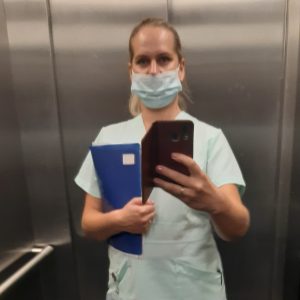 Lifte stovi šviesiai mėlynais chalatais ir veido kauke apsirengęs asmuo, kuris laiko mėlyną aplanką ir išmaniuoju telefonu daro asmenukę. Fone - metalinės lifto sienos.