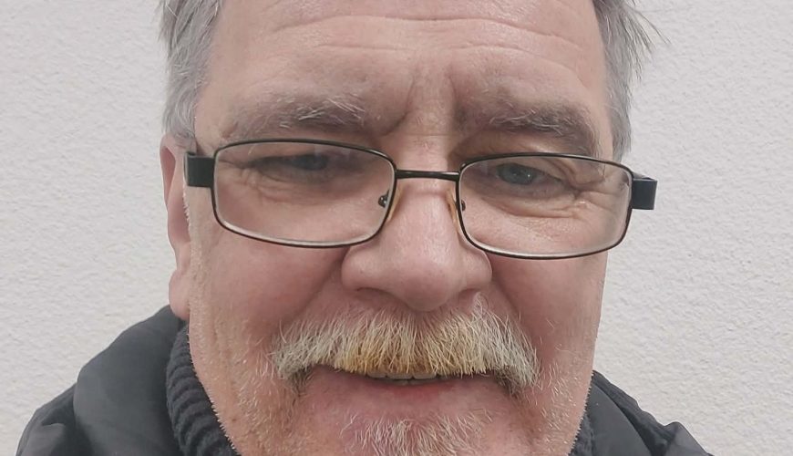 Vidutinio amžiaus vyras žilais plaukais, ūseliais ir akiniais šypsosi į kamerą. Jis vilki juodą megztinį su golfu ir juodą striukę, stovi priešais paprastą baltą faktūrinę sieną.