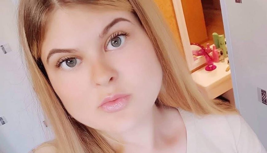Jauna moteris ilgais šviesiais plaukais ir šviesiomis akimis daro asmenukę patalpoje. Ji vilki šviesią palaidinę ir stovi priešais vonios kambarį su atviromis durimis ir matoma kriaukle.