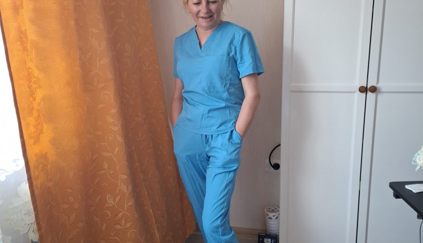 Moteris, vilkinti šviesiai mėlynus medicininius chalatus ir avinti baltas klumpes, stovi prie lango su oranžine užuolaida ir šypsosi susikišusi rankas į kišenes. Fone matoma balta spinta ir dalis stalo.
