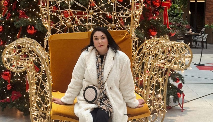 Asmuo baltu paltu ir languotu šaliku sėdi ant didelės, auksinės spalvos, šviesomis dengtos kėdės priešais papuoštą eglutę su raudonais ornamentais šventiškoje patalpos aplinkoje.