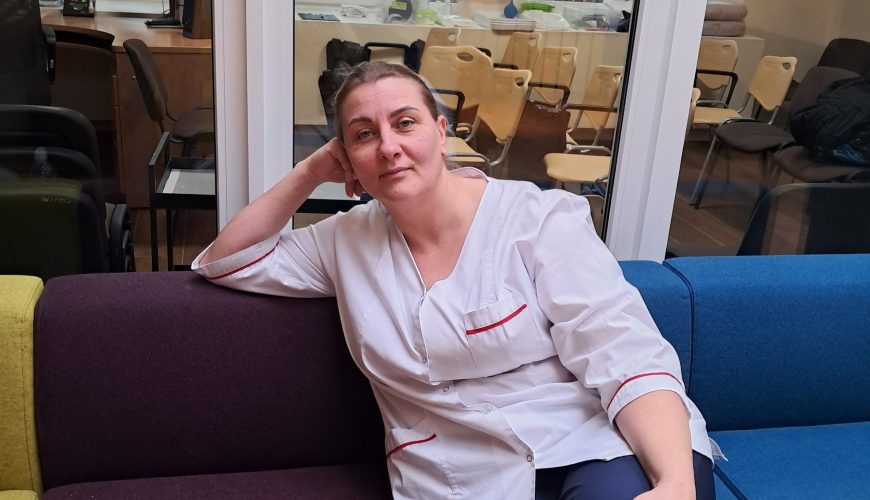 Moteris baltu chalatu sėdi ant spalvingos sofos, padėjusi galvą ant rankos. Ji yra patalpoje, o už jos esanti stiklinė siena rodo kitą kambarį su lauke matomais stalais, kėdėmis ir medžiais.