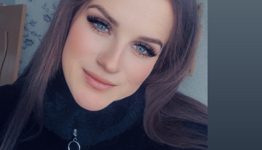 A woman with long brown hair and blue eyes smiles softly at the camera. She is wearing a black fuzzy jacket and is indoors with light-colored walls and patterned wallpaper in the background.