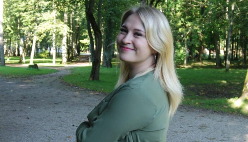 Besišypsanti moteris ilgais šviesiais plaukais, vilkinti žalią palaidinę ir džinsus, stovi sukryžiavusi rankas ant žvyruoto tako saulės apšviestame, žaliame parke, apsuptame medžių.