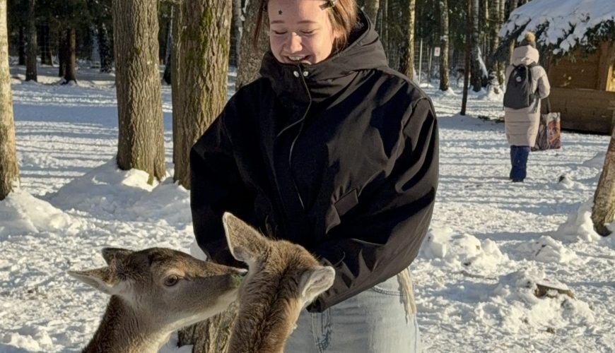 Juodą striukę ir šviesius džinsus vilkintis asmuo stovi snieguotame miške ir šypsodamasis šeria tris elnius. Saulės spinduliai apšviečia apsnigtą žemę ir medžius. Fone matomas dar vienas einantis asmuo.