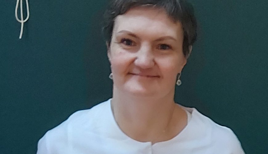 A woman with short brown hair, wearing a white top, stands in front of a dark green background, smiling softly at the camera.