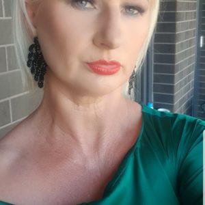 A woman with short blonde hair wearing black earrings and a green top looks directly at the camera, standing in front of a brick wall.