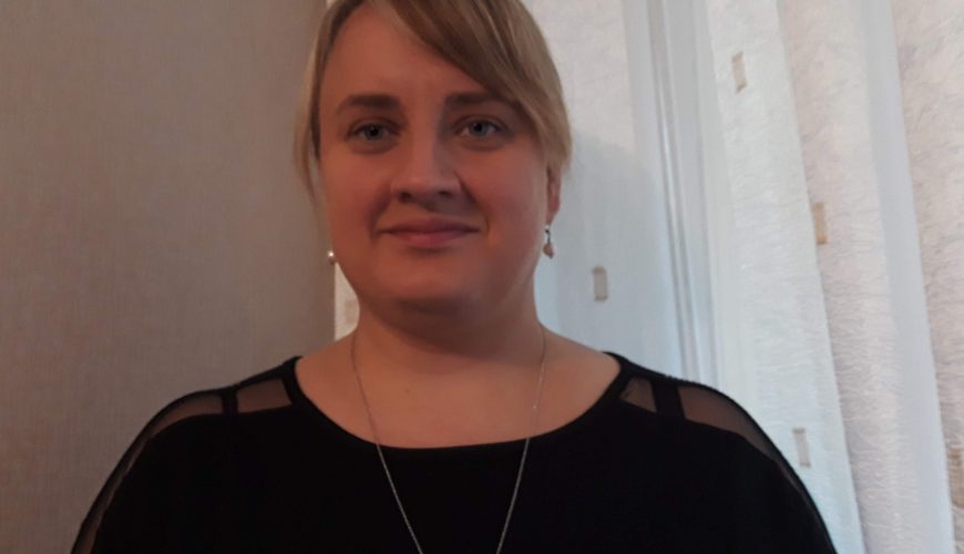 Moteris šviesiais plaukais, surištais atgal, vilkinti juodą liemenėlę, auskarus su kabliuku ir grandinėlę su pakabuku, stovi patalpoje priešais baltas užuolaidas.