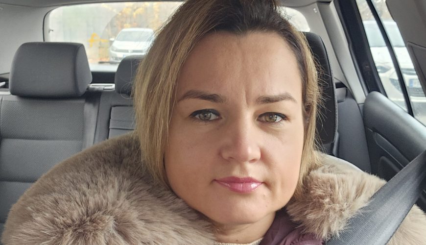 Automobilyje sėdi moteris trumpais šviesiais plaukais ir rimta veido išraiška, vilkinti violetinės spalvos paltą su didele kailine apykakle ir prisisegusi saugos diržą. Fone matomas automobilio salonas ir stovinčios transporto priemonės.