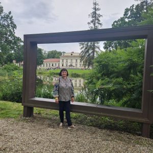 A woman stands on gravel in front of a large, rectangular wooden frame with a bench, with a pond, trees, and a historic building in the background.