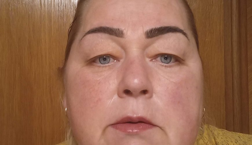 Moteris šviesia oda, mėlynomis akimis ir tamsiais ryškiais antakiais žiūri tiesiai į kamerą. Jos šviesūs plaukai sušukuoti atgal, ji vilki geltoną palaidinę. Fone matomos medinės durys.