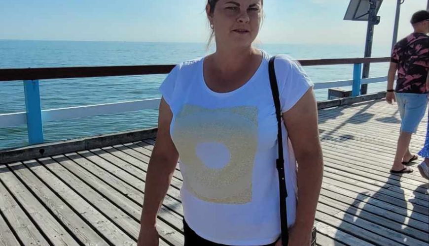 Saulėtą dieną moteris, vilkinti baltus marškinėlius, juodus šortus ir avinti baltus sportbačius, eina mediniu molu prie jūros. Fone mėlynas dangus ir ramus vanduo.