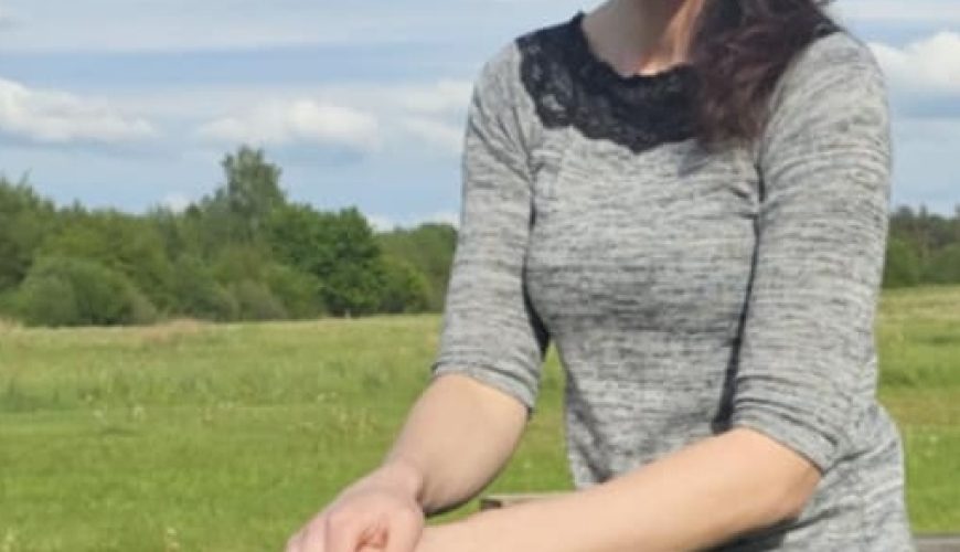 Saulėtą dieną su mėlynu dangumi ir išsibarsčiusiais debesimis moteris ilgais rudais plaukais, vilkinti pilką palaidinę ir juodas kelnes, sėdi ant medinio suoliuko žolėtame lauke ir šypsosi į kamerą.