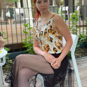 A woman with red hair sits on a white chair outdoors. She is wearing a sleeveless animal print top and a black sheer skirt. She looks directly at the camera with a neutral expression. Green plants and a black fence are in the background.