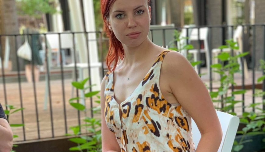 Moteris raudonais plaukais sėdi ant baltos kėdės lauke, vilkėdama gyvūnų raštais margintą palaidinę be rankovių ir juodą permatomą sijoną. Ji žiūri tiesiai į kamerą, o fone matoma žaluma ir medinis paklotas.