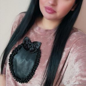 A woman with long black hair and bold makeup wears a light pink velvet top with a large black decorative patch framed by beads. She is standing indoors against a plain light background.
