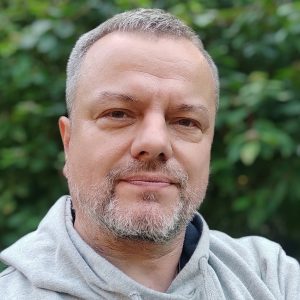 Vidutinio amžiaus vyras trumpais žilais plaukais ir barzda, vilkintis šviesiai pilką gobtuvą su gobtuvu, stovi lauke priešais žalią lapiją ir neutraliai žiūri į kamerą.
