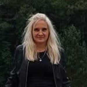 Moteris ilgais šviesiais plaukais, vilkinti juodą palaidinę ir juodą striukę, stovi lauke priešais žalius medžius ir šiek tiek šypsosi į kamerą.