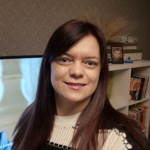Moteris tiesiais rudais plaukais ir baltu raštuotu megztiniu šypsosi į kamerą. Už jos stovi lentyna su knygomis, įrėminta nuotrauka ir televizoriaus ekranas, kuriame rodomas neryškus vaizdas.