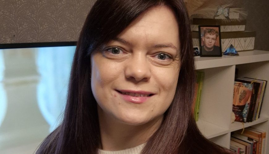 Moteris tiesiais rudais plaukais ir baltu raštuotu megztiniu šypsosi į kamerą. Už jos stovi lentyna su knygomis, įrėminta nuotrauka ir televizoriaus ekranas, kuriame rodomas neryškus vaizdas.