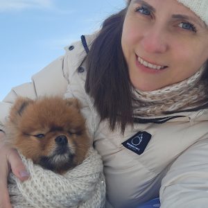 A woman in a white winter coat, knit scarf, and hat smiles while holding a small fluffy dog wrapped in a matching knit blanket. The background is a bright blue sky.