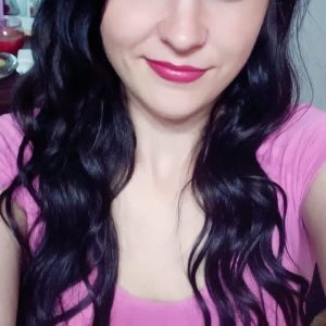 A woman with long wavy dark hair and blue eyes is smiling at the camera. She is wearing a pink top and is indoors, with some household items visible in the background.
