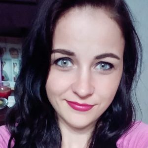 Į kamerą šypsosi ilgų, banguojančių tamsių plaukų ir mėlynų akių moteris. Ji vilki rausvą palaidinę ir atrodo esanti patalpoje.