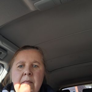 Šviesiai rudų plaukų ir mėlynų akių moteris sėdi automobilyje ir žiūri į kamerą. Saulės šviesa meta šešėlį ant jos veido, o virš jos ir už jos matomas automobilio stogas ir langai.