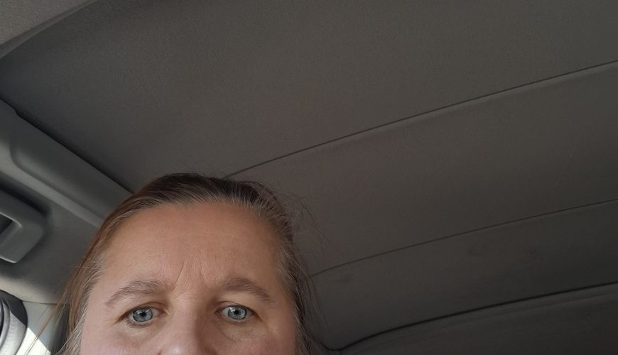 Šviesiai rudų plaukų ir mėlynų akių moteris sėdi automobilyje ir žiūri į kamerą. Saulės šviesa meta šešėlį ant jos veido, o virš jos ir už jos matomas automobilio stogas ir langai.
