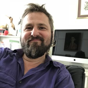 Violetiniais marškiniais vilkintis barzdotas vyras šypsosi į kamerą sėdėdamas patalpoje, o fone matomas "Apple iMac" kompiuteris ir įrėmintas paveikslas.