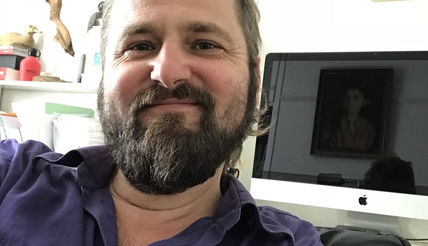 Violetiniais marškiniais vilkintis barzdotas vyras šypsosi į kamerą sėdėdamas patalpoje, o fone matomas "Apple iMac" kompiuteris ir įrėmintas paveikslas.