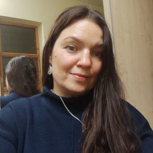 Į kamerą šypsosi moteris ilgais rudais plaukais, vilkinti tamsiai mėlyną palaidinę, sidabrinę grandinėlę ir auskarus. Už jos stovi veidrodis, kuriame atsispindi dalis jos nugaros ir plaukų.