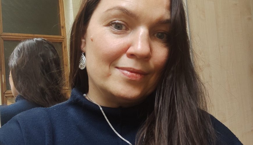 Į kamerą šypsosi moteris ilgais rudais plaukais, vilkinti tamsiai mėlyną palaidinę, sidabrinę grandinėlę ir auskarus. Už jos stovi veidrodis, kuriame atsispindi dalis jos nugaros ir plaukų.