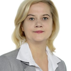 Šviesiaplaukė moteris, vilkinti šviesiai pilką švarką ir baltą marškinių su apykakle, pozuoja prieš paprastą baltą foną, neutraliu veidu žiūrėdama tiesiai į fotoaparatą.