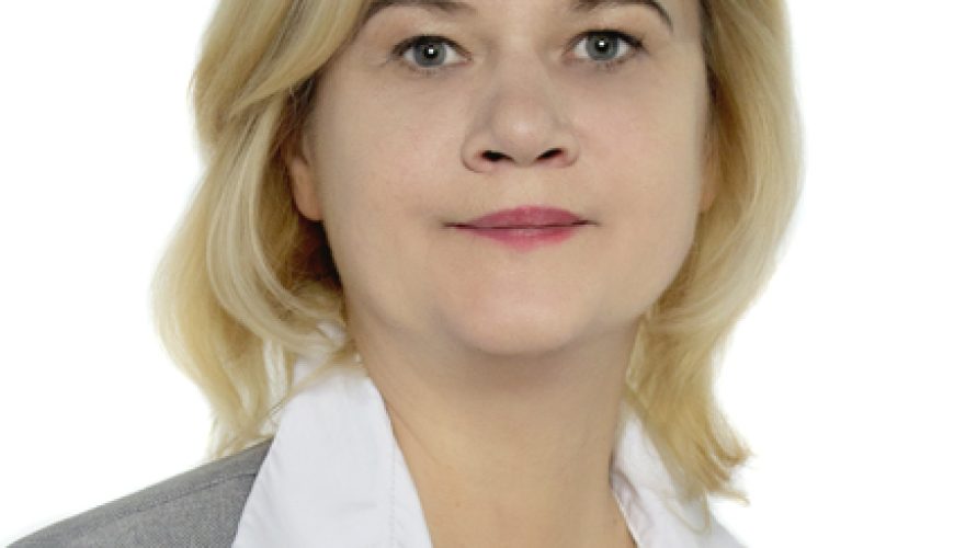 Šviesiaplaukė moteris, vilkinti šviesiai pilką švarką ir baltą marškinių su apykakle, pozuoja prieš paprastą baltą foną, neutraliu veidu žiūrėdama tiesiai į fotoaparatą.
