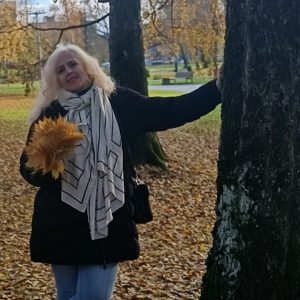 Parke prie medžio stovi šviesiaplaukė moteris, apsirengusi juodu paltu, šviesiu šaliku ir mėlynais džinsais. Ji laiko puokštę geltonų rudens lapų, o ant žemės guli nukritę lapai.