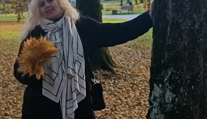 Parke prie medžio stovi šviesiaplaukė moteris, apsirengusi juodu paltu, šviesiu šaliku ir mėlynais džinsais. Ji laiko puokštę geltonų rudens lapų, o ant žemės guli nukritę lapai.