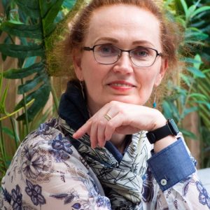 Moteris su garbanotais raudonais plaukais, akiniais ir gėlių raštuota palaidine sėdi priešais žalius augalus, atsirėmusi smakru į ranką ir švelniai šypsosi. Ji dėvi skaraitę ir išmanųjį laikrodį.