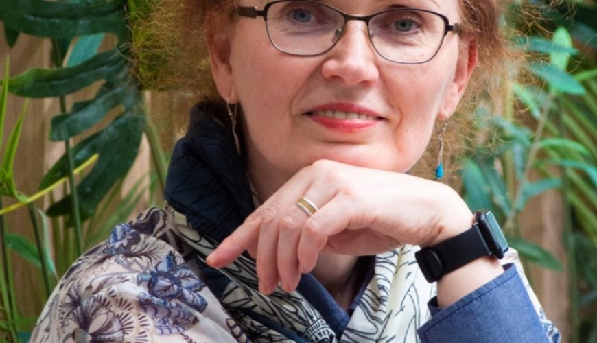 Moteris su garbanotais raudonais plaukais, akiniais ir gėlių raštuota palaidine sėdi priešais žalius augalus, atsirėmusi smakru į ranką ir švelniai šypsosi. Ji dėvi skaraitę ir išmanųjį laikrodį.
