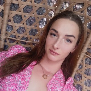 Moteris su ilgais rudais plaukais, vilkinti rožinį marškinėlį su gėlių motyvais ir apvalią pakabuką ant kaklo, atsipalaidavusi sėdi ant pintos kėdės ir žiūri į fotoaparatą.