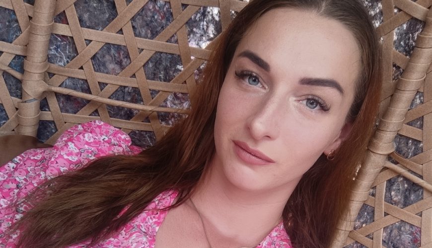 Moteris su ilgais rudais plaukais, vilkinti rožinį marškinėlį su gėlių motyvais ir apvalią pakabuką ant kaklo, atsipalaidavusi sėdi ant pintos kėdės ir žiūri į fotoaparatą.